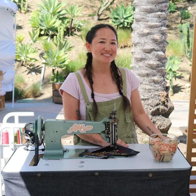 A woman stands behind a table at an outdoor market under a white canopy, wearing a green apron over a light pink shirt. On the table is a vintage-style teal chainstitch sewing machine and a denim tablecloth with the brand name 'Aki&co' in white script.