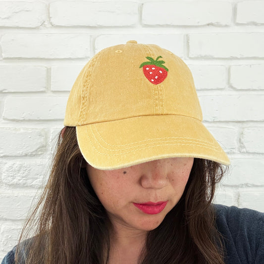Yellow cap with strawberry design worn by a person against a white brick wall.